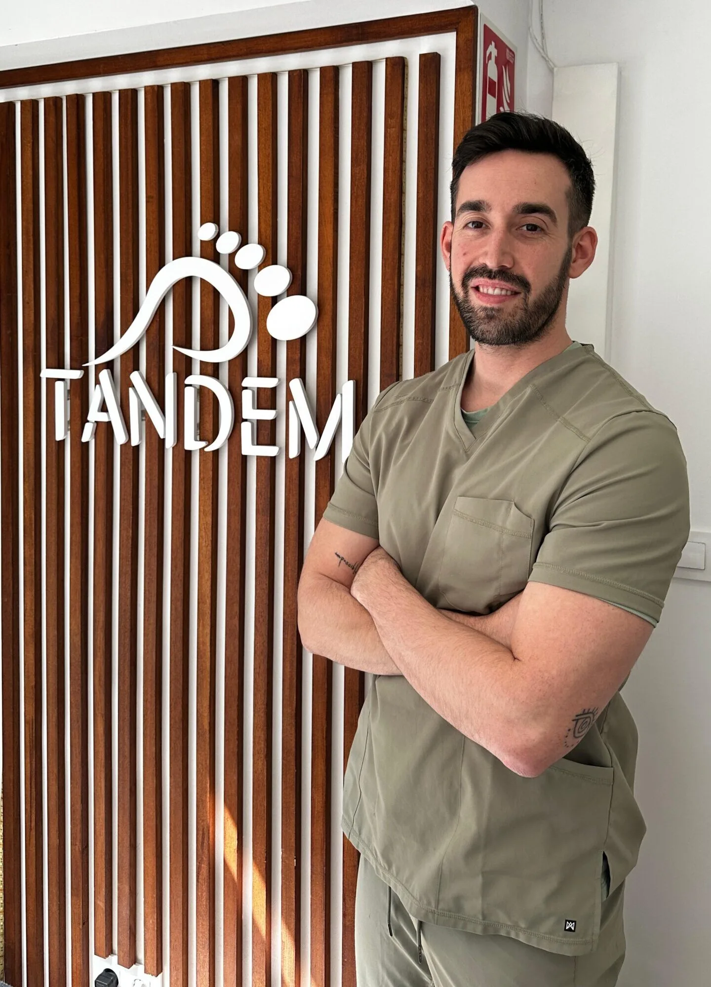 Podólogo con uniforme verde oliva posando frente al logotipo de la clínica Tándem Podología